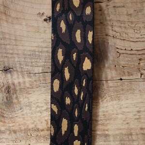 Peut inclure: Une sangle en tissu tissé présentant un motif léopard répétitif dans des tons de noir, brun foncé et beige. Le matériau présente un tissage texturé visible, créant un motif de surface cohérent et dense sur toute la longueur de la sangle.