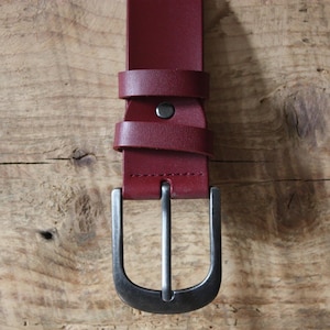 Op de afbeelding: Een diepbordeauxrode riem met een gladde, mat afgewerkte band en een donkere gesp van geborsteld metaal. De riem is voorzien van twee bijpassende bordeauxrode lederen lussen en een enkele metalen klinknagel die de band bij de gesp vastzet.