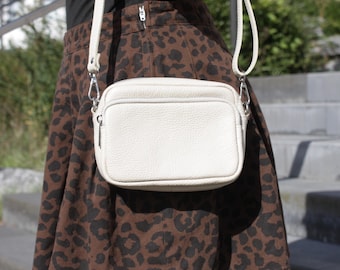 Puede incluir: Un bolso bandolera rectangular de color blanco roto con textura granulada, que cuenta con un cierre de cremallera superior y una correa de hombro ajustable a juego sujeta con herrajes metálicos plateados, sobre una falda con estampado de leopardo.