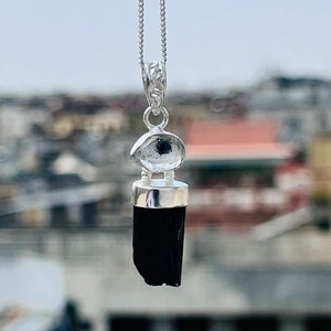 May include: A silver pendant necklace featuring a black tourmaline crystal with a clear quartz accent. The pendant hangs from a delicate chain. The necklace is displayed against a blurred outdoor background.