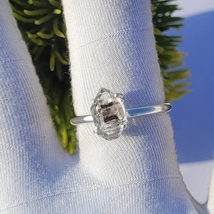 May include: A silver ring featuring a rough-cut, clear crystal gemstone with dark inclusions. The ring is set on a simple silver band, showcasing the unique shape and texture of the stone. The ring is displayed on a white fabric surface.