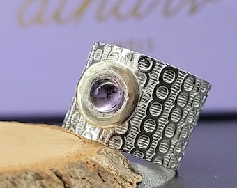 May include: A silver ring with a textured surface and a round, purple gemstone set in a gold bezel. The ring is on a wooden surface.