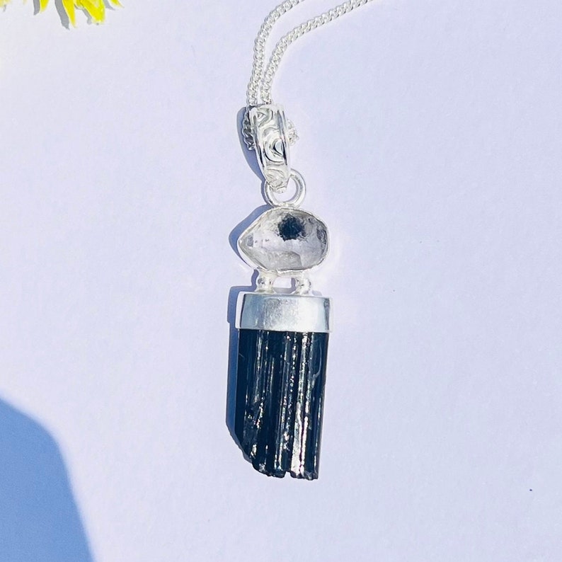 May include: A silver pendant necklace featuring a black tourmaline crystal and a clear quartz stone. The pendant hangs from a delicate silver chain. The black tourmaline is a dark, elongated shape, and the quartz is a rounded, faceted stone.