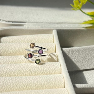 Op de afbeelding: Een zilverkleurige ring met vier gekleurde edelstenen: amber, paars, rood en groen. De ring heeft een uniek, wervelend ontwerp, waarbij elke edelsteen op een aparte band is gezet. De ring wordt gepresenteerd op een getextureerd, gebroken witte ondergrond.