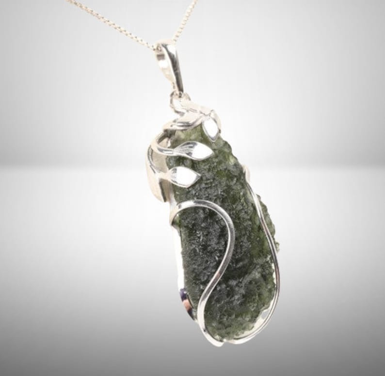 Pode incluir: Um pingente de prata com uma pedra de moldavite verde engastada em um delicado design de folhas de prata.