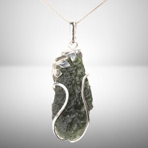 Pode incluir: Um pingente de prata com uma pedra de moldavite verde engastada em um delicado design floral de prata.