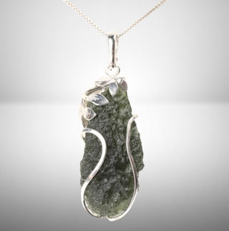 Pode incluir: Um pingente de prata com uma pedra de moldavite verde engastada em um delicado design floral de prata.