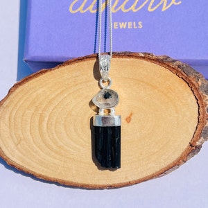 May include: A silver pendant necklace featuring a black tourmaline crystal and a clear quartz stone. The pendant hangs from a delicate silver chain. The necklace is displayed on a wooden surface.