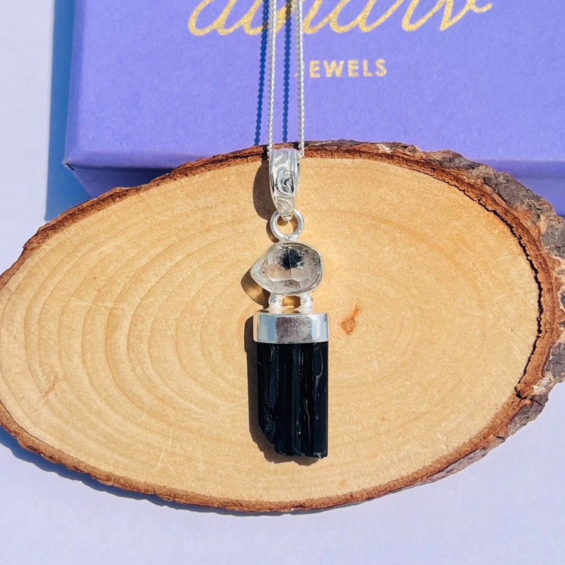 May include: A silver pendant necklace featuring a black tourmaline crystal and a clear quartz stone. The pendant hangs from a delicate silver chain. The necklace is displayed on a wooden surface.