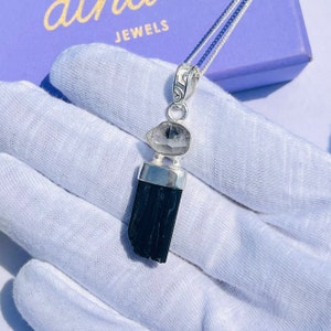 May include: A silver pendant necklace featuring a black tourmaline crystal and a clear quartz stone. The pendant hangs from a delicate silver chain. The necklace is displayed on a white surface, with the brand name "althea JEWELS" visible in the background.