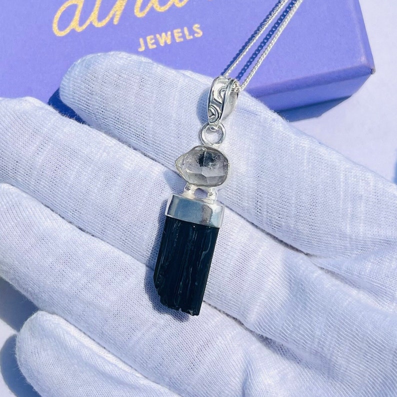 May include: A silver pendant necklace featuring a black tourmaline crystal and a clear quartz stone. The pendant hangs from a delicate silver chain. The necklace is displayed on a white surface, with the brand name "althea JEWELS" visible in the background.