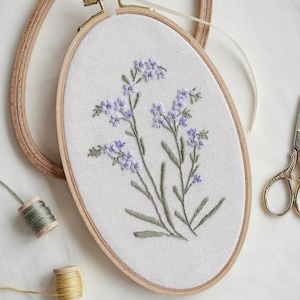 May include: A close-up of a hand-stitched embroidery hoop with a floral design. The hoop is made of wood and has a white fabric background. The embroidery features delicate purple flowers and green leaves.