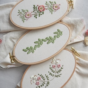 May include: Three oval embroidery hoops with different floral designs. The top hoop features pink flowers and green leaves, the middle hoop has green leaves, and the bottom hoop has pink and white flowers with green leaves.