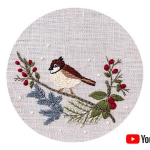 Puede incluir: Diseño bordado de un pájaro marrón posado en una rama con hojas verdes y azules y bayas rojas. El fondo es una tela de lino beige claro.