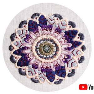 May include: A circular embroidery design featuring a mandala with intricate details in shades of purple, blue, grey, and pink. The mandala is surrounded by a border of smaller, repeating patterns.