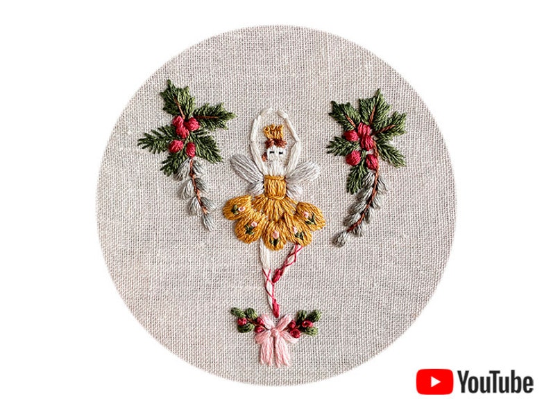 May include: Embroidered hoop art featuring a yellow and white ballerina with a red bow, surrounded by green holly sprigs with red berries.