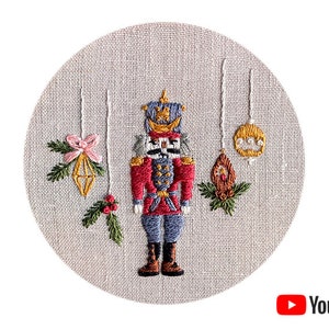 May include: Embroidered nutcracker with a red and blue uniform, holding a sword, surrounded by three hanging ornaments with gold, pink, and brown details. The background is a beige linen fabric.