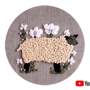 Puede incluir: Arte de bordado en aro con una oveja blanca con patas y cara negras, rodeada de delicadas flores blancas con centros azul claro sobre un fondo de lino gris.