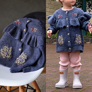 Op de afbeelding: Marineblau vest met bloemenborduurwerk en houten knopen. Het vest heeft een gerimpeld ontwerp. De afbeelding toont het vest op een witte stoel en gedragen door een kind met roze leggings en witte laarzen.