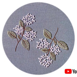 Puede incluir: Flores de lila bordadas sobre un fondo de lino gris. Las flores son en tonos de morado y blanco, con hojas verdes.