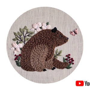 May include: Embroidered brown bear sitting in a circle of pink and white flowers and green leaves. A small pink butterfly is flying to the right of the bear.