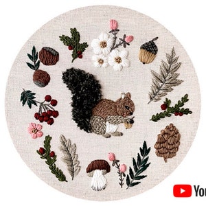 May include: Embroidered hoop art featuring a brown squirrel holding an acorn, surrounded by various autumnal elements like leaves, berries, and mushrooms. The background is a beige fabric.