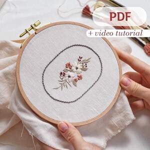 May include: A close-up of a hand holding a wooden embroidery hoop with a floral design embroidered on white fabric. The design features a cluster of small white and pink flowers with green leaves, all enclosed in a grey oval. The text "PDF + video tutorial" is visible in the top right corner of the image.
