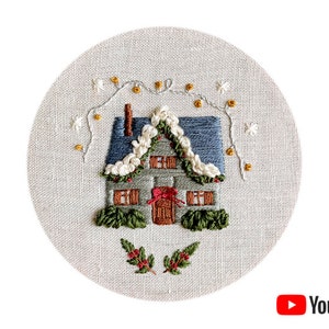 Puede incluir: Diseño bordado de una pequeña casa con un techo blanco nevado, arbustos verdes y un lazo rojo en la puerta. La casa está rodeada de una guirnalda de flores blancas y luces doradas sobre un fondo de tela beige.