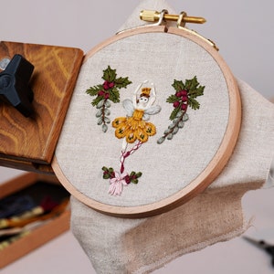 May include: A wooden embroidery hoop with a beige linen fabric stretched across it. The fabric features an embroidered design of a ballerina in a yellow dress with red berries and green leaves. The hoop is attached to a wooden stand with a black knob.