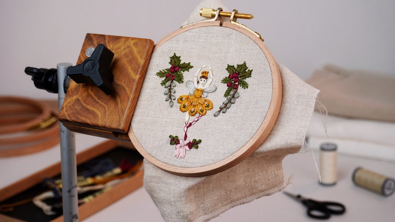 May include: A wooden embroidery hoop with a beige linen fabric stretched across it. The fabric features an embroidered design of a ballerina in a yellow dress with red berries and green leaves. The hoop is attached to a wooden stand with a black knob.