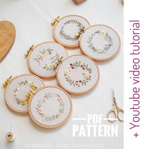 May include: Five embroidery hoop patterns featuring floral wreaths with different colored flowers and leaves. The hoops are on a white surface with a pair of scissors and a spool of thread. The text "PDF PATTERN" is written on the image. The text "+ Youtube video tutorial" is written on the right side of the image.