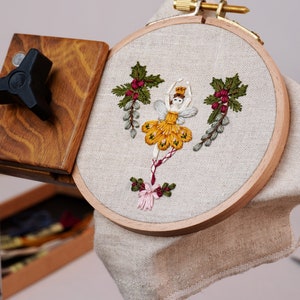 May include: A close-up of a hoop with a white fabric stretched across it. The fabric is embroidered with a yellow and green fairy figure with red berries and green leaves. The hoop is attached to a wooden stand.