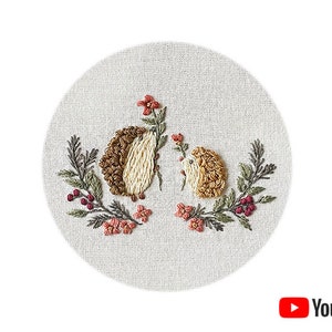 Pode incluir: Desenho bordado de dois ouriços com pelo castanho e branco, rodeados de folhas verdes e flores cor-de-rosa sobre um fundo de tecido bege.