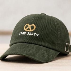 May include: An olive green corduroy baseball cap featuring an embroidered golden pretzel icon above the white text "STAY SALTY". The cap includes a curved brim, stitched eyelets, and a silver-toned metal adjustable side buckle strap.