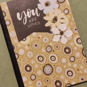 May include: A decorative notebook with a patterned cover featuring a repeating design of circles in shades of brown and cream. The cover includes a black banner with the words "you are loved" in white script, and three paper flowers with glitter centers.