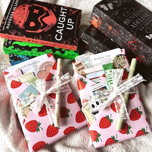 Op de afbeelding: Twee ingepakte cadeautjes met een roze aardbeienpatroon, vastgebonden met wit kanten lint en een lichtgekleurde pen. Elk pakketje bevat briefpapier zoals een leeslogboek en stickers. Daarachter liggen boeken met de titels Caught Up, Game On en Hunting Adeline.