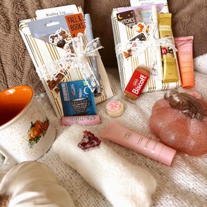 May include: Two gift packages wrapped in white paper with gold stripes, tied with white lace ribbon. Each contains a reading log, tea, snacks, and stationery. Includes a ceramic mug with a pumpkin print, a pink hand cream, and a fuzzy white sock.