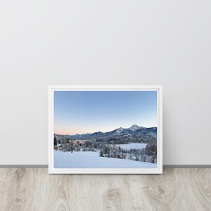 Puede incluir: Una fotografía enmarcada de un paisaje montañoso cubierto de nieve bajo un cielo azul claro. La fotografía, en un marco blanco, representa una serena escena invernal con árboles cargados de nieve y un lago tranquilo. La paleta de colores general está dominada por blancos, azules y toques de rosa.