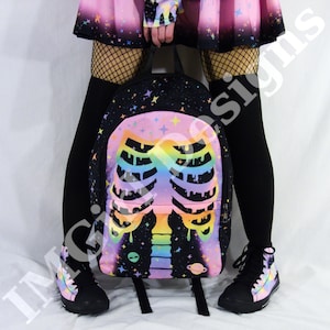 May include: A black backpack featuring a colourful rainbow gradient ribcage graphic with a dripping effect. The design includes small stars, a planet, and an alien head against a dark space background. The backpack has black straps and zippers.