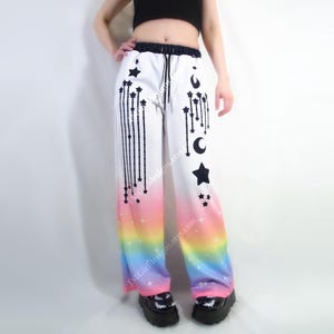 May include: White palazzo pants with a celestial design. The pants feature black shooting stars, moons, and stars, with a rainbow gradient at the bottom. The pants have a drawstring waist and are wide-legged.