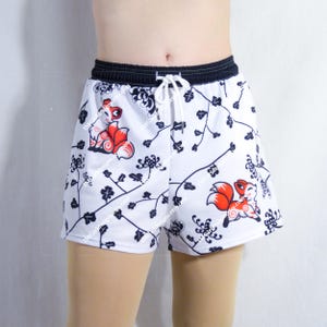 May include: White athletic shorts featuring a repeating pattern of dark blue floral vines and orange and white fox illustrations. The shorts have a dark blue elastic waistband with a white drawstring cord tied at the center front for adjustment.