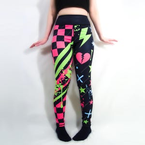 Könnte beinhalten: Leggings mit einem auffälligen, geteilten Design. Eine Seite zeigt ein rosa-schwarzes Schachbrettmuster mit neongrünen Streifen und Farbspritzern. Die andere Seite hat einen schwarzen Hintergrund mit einem gebrochenen Herzen, Sternen und Blitzen.