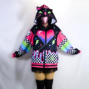 Puede incluir: Una sudadera con capucha negra con cremallera y capucha con orejas de gato, con un corazón pixelado, rayos y un diseño de pintura goteando. Las mangas tienen un patrón de cuadros y detalles de arcoíris. La parte inferior tiene flecos negros.