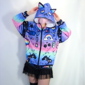 May include: A blue, pink, and purple zip-up hoodie with a pixelated video game design. The hoodie features a cat ear hood, a rainbow, and controller graphics. The text "GAME OVER" and "LIFE UVEK" are visible on the hoodie.