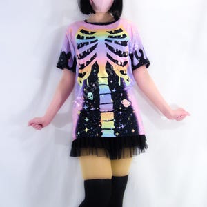 May include: A short-sleeved, colorful t-shirt dress with a skeleton rib cage design. The design features a rainbow-colored rib cage over a galaxy-themed background with stars, planets, and a black tulle skirt.
