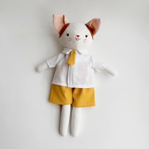 May include: A white stuffed cat doll wearing a white shirt with a yellow tie and yellow shorts. The doll has brown ears and a pink nose.