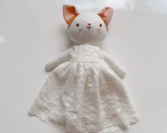 May include: White stuffed cat doll wearing a white lace dress with a brown and orange bow on its head.