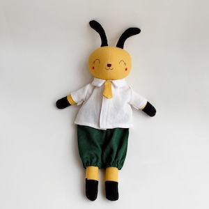 Peut inclure: Un jouet en peluche jaune avec des oreilles noires, une chemise blanche et un pantalon vert. Le jouet a un visage souriant avec des joues rouges et un nez brun.