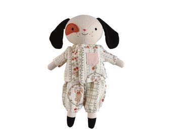 Puede incluir: Un muñeco de perro de peluche con cabeza color crema, orejas negras y una mancha roja alrededor de un ojo. El muñeco lleva un conjunto de pijama estampado con un bolsillo rosa. El pijama tiene un diseño floral y a cuadros.