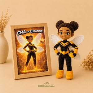 May include: A crochet doll featuring a character with dark brown hair in two buns, wearing a black and yellow superhero suit with white wings. Beside it is a framed print showing the same character with the text "CHA of CHANGE" above.
